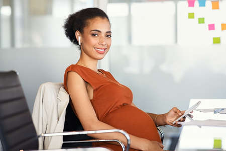Portrait of smiling pregnant business woman using smartphone and doing video call with wireless earphone in modern office. Formal pregnant black businesswoman texting on phone in office while looking at camera. Portrait of happy african american mom expecting baby sitting in meeting room.の写真素材