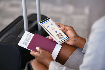 Close up hand of black businesswoman checking flight eticket on phone while holding passport and boarding pass. African american woman hand checking online travel plane ticket on mobile phone. Detail of business woman with flight ticket in waiting room at airport.の写真素材