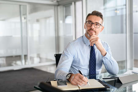 Thoughtful mature business man taking notes in notebook while using digital tablet at office. Mid adult businessman entrepreneur writing details on book while working in modern office. Pensive businessman with spectacles planning with copy space.の写真素材