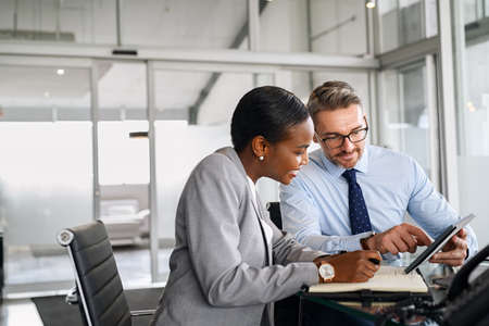 Mature businessman discussing work with black manager writing notes in diary. Confident business man in formal clothing working with african american woman showing data on digital tablet. Smiling professional business people working together on new business strategy.の写真素材
