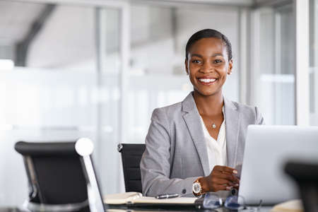 Portrait of a cheerful businesswoman sitting at desk in modern office and looking at camera. Attractive and successful black business woman sitting at conference table with laptop. Smiling african american executive using laptop while working from office with copy space.の写真素材