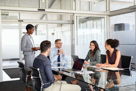 Smiling business team discussing work in meeting using laptop and documents in conference room. Successful business woman and businessmen discussing marketing strategy and ideas at meeting room. Corporate business man and woman with manager and secretary working together.の写真素材