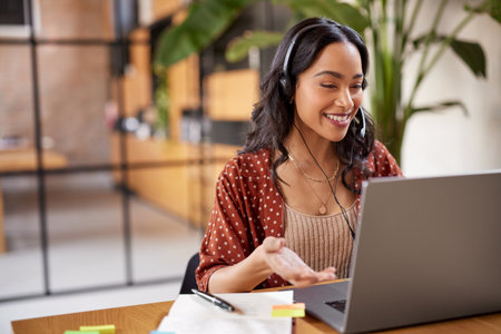 Smiling successful business woman with headphones talking while attending a meeting online using laptop. Latin woman using computer with headset and microphone in an online meeting. Hispanic businesswoman watch webinar and listen online course.の写真素材
