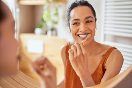 Smiling young woman brushing teeth in bathroom. Happy girl looking in mirror while using ecological toothbrush with whitening toothpaste to clean her teeth. Multiethnic girl cleaning teeth in the morning time.の写真素材