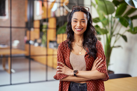Portrait of smiling young multiethnic woman looking at camera with crossed arms. Successful latin business woman standing in modern office with copy space. Young university hispanic girl with pleasant smile and folded arms.の写真素材