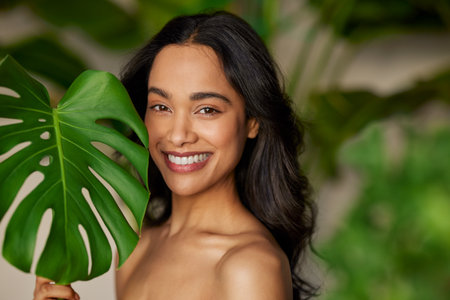 Close up face of beautiful young multiethnic woman covering her face with green monstera leaf while looking at camera. Portrait of beauty girl with natural makeup standing behind big green leaves with copy space. Portrait of beautiful mixed race woman in nature.の写真素材
