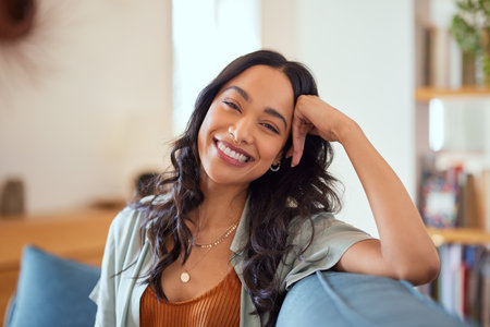 Portrait of happy young latin woman sitting on sofa at home while looking at camera. Happy multiethnic carefree woman in casual clothing relaxing at home sitting on couch. Cheerful mexican girl sitting on couch and laughing.の写真素材