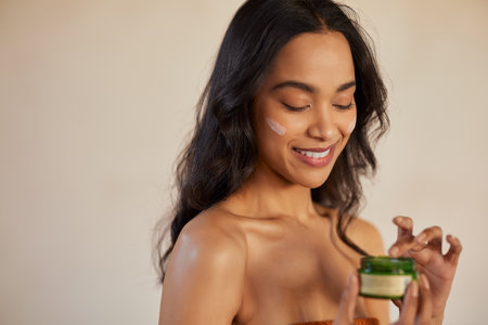 Girl caring of her beautiful skin face isolated on beige background. Young multiethnic woman applying eco friendly moisturizing cream on face with copy space. Smiling natural mixed race girl holding little green jar of ecological cosmetic moisturizer while applying cream on cheek.の写真素材