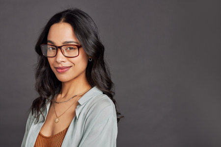 Confident young multuethnic woman wearing eyeglasses and standing on gray wall with copy space. Portrait of smiling business woman isolated against gray wall while looking at camera. Proud and successful university student girl with specs.の写真素材