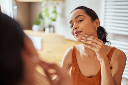 Pretty young woman touching and examining her face skin while looking in mirror in the morning. Beautiful latin woman enjoying hydrated skin in her bathroom. Happy self loving girl enjoying her natural beauty complexion, during morning beauty routine.の写真素材