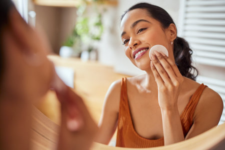 Portrait of latin young beauty woman looking at mirror while cleaning her face with cotton disc in bathroom. Beautiful multiethnic woman taking good care of her face while standing against mirror in bathroom. Enjoying facial skincare routine concept.の写真素材
