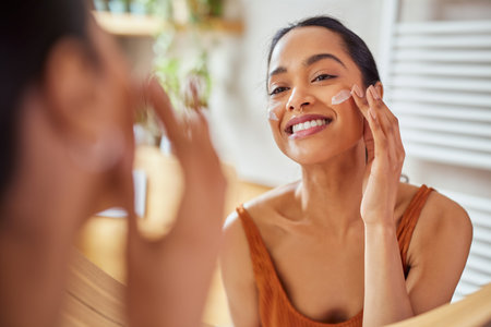 Portrait of smiling latin woman applying cream on her face while looking in mirror in her bathroom. Young multiethnic woman applying moisturizer on her cheek while looking in the mirror. Happy mexican girl using lotion every morning.の写真素材