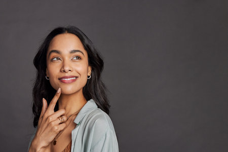 Portrait of pensive young mixed race woman with hand on chin looking away isolated on gray background. Multiethnic beautiful girl in casual clothing isolated against gray wall daydreaming with copy space. Close up face of happy mexican woman thinking and making plans about future.の写真素材
