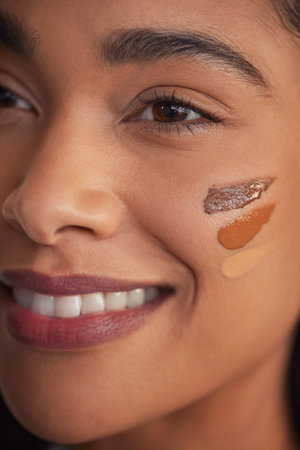 Closeup portrait of young smiling woman applying foundation swatches of various tones on face. Beautiful hispanic girl with different shades of liquid concealer on cheek. Detail of multiethnic woman trying different shades of foundation on skin while looking away.の写真素材