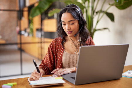 Successful young business woman working on laptop with headphones in a call center as a consultant. Happy businesswoman with headset translating and writing notes by listening to audio course. University girl listening online lesson on laptop and writing notes at home while studying.の写真素材