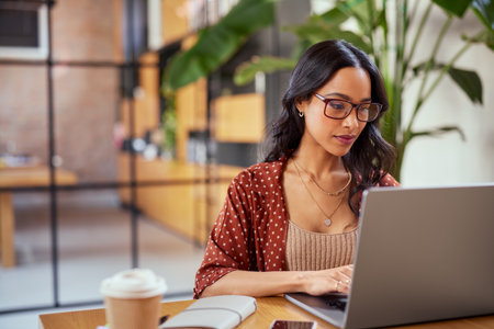 Concentrated latin business woman working on laptop wearing eyeglasses. Focused woman entrepreneur working on laptop in office lobby. Young serious multiethnic businesswoman working online on computer wearing spectacles.の写真素材