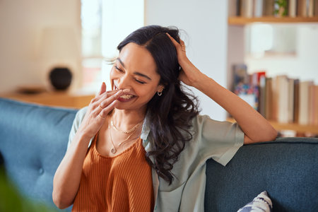 Happy young latin woman sitting on sofa at home and laughing. Happy multiethnic carefree woman in casual clothing relaxing at home while laughing with hand near mouth. Close up face of cheerful girl sitting on couch and laughing while thinking of something funny.の写真素材