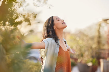 Young latin woman with arms outstretched breathing in fresh air during sunrise at the balcony. Healthy girl enjoying nature while meditating during morning with open arms and closed eyes. Mindful woman enjoying morning ritual while relaxing in outdoor park.の写真素材