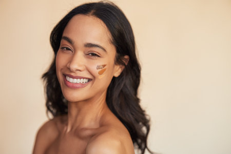Portrait of young mixed race smiling woman applying foundation swatches of various tone on cheek. Beautiful hispanic woman with different shades of foundation on face isolated against background with copy space. Attractive multiethnic girl trying concealer shades while looking at camera.の写真素材