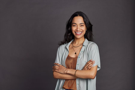 Portrait of smiling young multiethnic woman in casual clothing with arms crossed standing against gray background. Cheerful latin woman with folded hands looking at camera with big grin. Satisfied mexican girl with copy space isolated against gray background.の写真素材