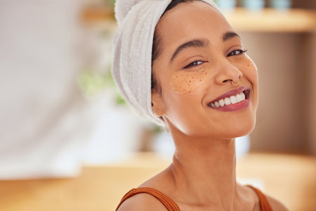 Portrait of latin woman with under eye collagen mask looking at camera with copy space. Healthy young woman taking care of her skin by applying under eye moisturizing mask patches. Mexican girl with towel on head after shower smiling with eye detox patches for puffiness and dark circles.の写真素材