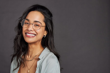 Casual cheerful young woman with eyeglasses smiling at camera on gray background. Close up face of happy multiethnic girl laughing with trendy eyeglasses and copy space. Beautiful mixed race girl with specs having fun isolated against gray wall.の写真素材