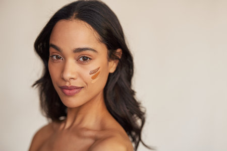 Portrait of young latin hispanic woman applying foundation swatches of various tone on face while looking at camera. Beautiful mixed race makeup artist with swatches of foundation cream on cheek isolated against beige background. Attractive young mexican woman with copy space looking at camera while trying concealer with different undertones.の写真素材