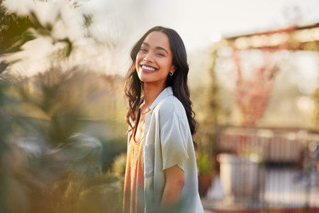 Young latin woman in casual clothing in the garden looking at camera, during early morning. Portrait of healthy mexican girl enjoying nature during sunset. Mindful multiethnic woman enjoy morning ritual with fresh air.の写真素材