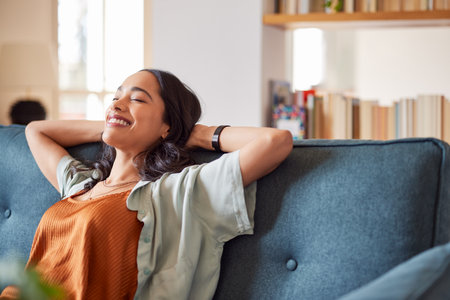Portrait of smiling young latin woman relaxing on couch with eyes closed. Serene young hispanic woman relaxing on sofa in living room with copy space. Happy beautiful multiethnic girl resting with hands behind head.の写真素材