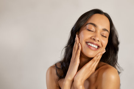 Portrait of beautiful latin woman touching her clean and healthy face against gray background. Smiling hispanic woman with natural makeup feeling healthy skin with eyes closed. Attractive multiethnic young woman pampering with perfect skin isolated with copy space.の写真素材