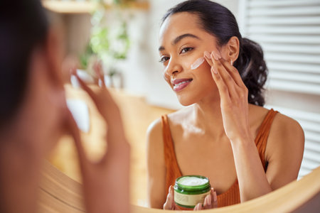 Woman caring of her beautiful skin on the face standing near mirror in the bathroom. Young mexican woman applying moisturizer on her face at home. Smiling multiethnic girl holding little green jar of bio skin cream and applying lotion on cheek.の写真素材