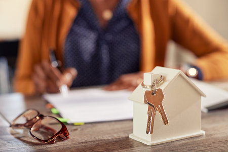 Successful independent african american woman in casual buying new home while filling out the sales documents in the background. Detail of keys from new home and tiny white model of house on work table of realtor with copy space.の写真素材