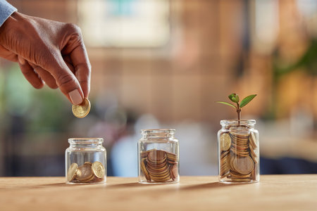 Close up of black woman hand adding money in coin in a jar. Girl hand holding coin adding money in glass jar of different sizes. Investment, savings and interest concept.の写真素材