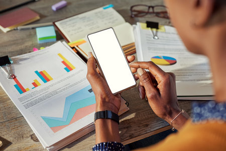 Mid adult woman using smartphone while working on project with graphs and reports in office. Close up of successful business black woman hands checking financial documents while using cellphone. Successful african american entrepreneur checking internet website at office desk with mobile.の写真素材