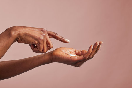 Elegant female hands with moisturizing cream closeup. Black woman applying organic cream on hands isolated on brown background. Close up shot of beautiful female hands holding and applying nourishing creamの写真素材