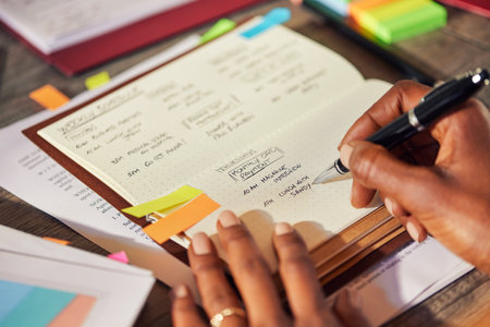 Closeup of african american woman hands making a list of work in diary on desk. Rear close up view of successful woman planning her week in advance while writing in personal organizer, using colored sticky notes. Detail of casual woman listing down tasks for coming week while working in office.の写真素材