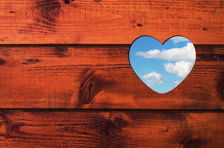 Pattern for text or card consists heart shaped hole with blue sky and white clouds in a brown wooden wall.の写真素材