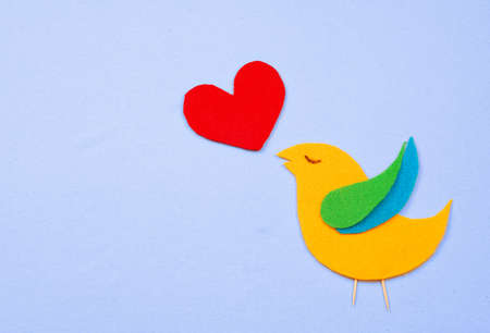 A whimsical, felt cutout bird shape in yellow, green and blue with a red felt heart floating that represent the bird's song. All are on a light blue cotton background. Copy space available.の写真素材