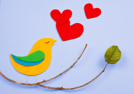 A whimsical, felt cutout bird shape in yellow, green and blue with red felt hearts floating that represent the bird's song. The bird is sitting on a twig with a single green silk leaf.の写真素材