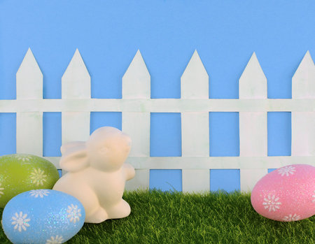 Cute Easter image with green grass, colorful sparkly eggs, a ceramic bunny and a white picket fence...