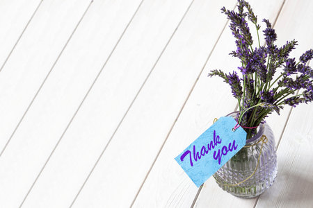 Bunch of lavender in a glass lantern jar,  with thank you written on a blue gift label.  White wood background