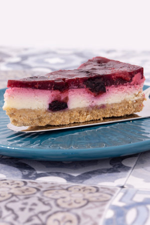 Slice of summer fruit cheesecake with a biscuit base served with a cake cutter utensil on a blue...