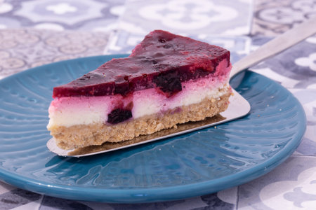 Slice of summer fruit cheesecake with a biscuit base served with a cake cutter utensil on a blue...