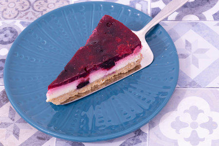 Slice of summer fruit cheesecake with a biscuit base served with a cake cutter utensil on a blue...