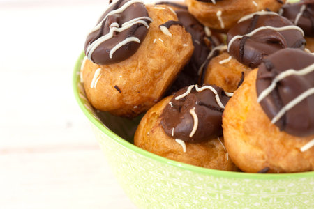 Profiteroles, coated in chocolate, stacked in a pattermed green and white bowl dish.  On a white...
