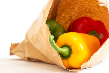 Yellow, red and green peppers in a brown paper bag on a white wood background.  Environmentally friendly packaging conceptの写真素材