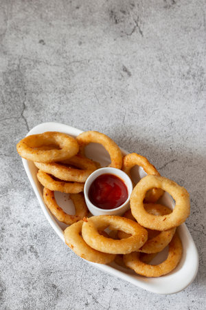 Battered onion rings in a dish,  with a tomato ketchup dip. On a concrete background