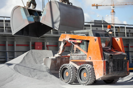 Crane loading cargo ship with gravel assisted by bobcat