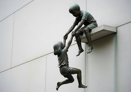 Bronze sculpture of a boy reaching down to help a girl climb onto a ledge.の素材