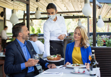 Handsome couple having lunch in the restaurant, being served by a waitress with a mask, coronavirus...